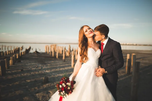 Vackra unga bröllopsparet, bruden och brudgummen poserar nära trästolpar i bakgrunden havet — Stockfoto