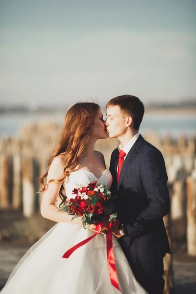 Casal de casamento jovem bonito, noiva e noivo posando perto de postes de madeira no fundo do mar — Fotografia de Stock