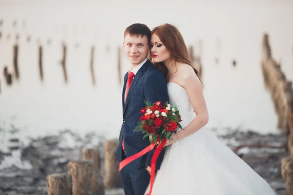 Schöne junge Hochzeitspaar, Braut und Bräutigam posiert in der Nähe von Holzpfählen auf dem Hintergrund Meer — Stockfoto