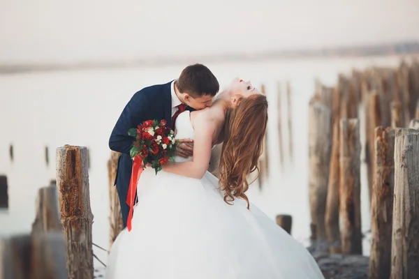 Casal de casamento, noivo, noiva com buquê posando perto do mar ao pôr do sol — Fotografia de Stock