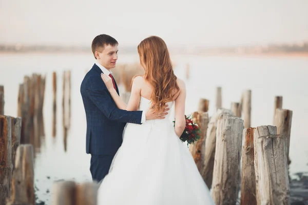 Casal de casamento, noivo, noiva com buquê posando perto do mar ao pôr do sol — Fotografia de Stock