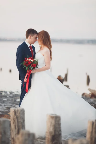 Casal de casamento jovem bonito, noiva e noivo posando perto de postes de madeira no fundo do mar — Fotografia de Stock