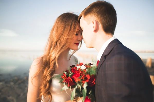 Wedding couple, groom, bride with bouquet posing near sea on sunset — Stock Photo, Image