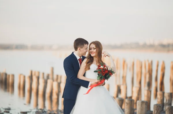 Casal de casamento jovem bonito, noiva e noivo posando perto de postes de madeira no fundo do mar — Fotografia de Stock