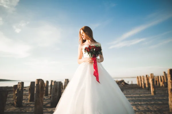 Hermosa novia romántica en vestido blanco posando sobre el fondo mar y postes de madera —  Fotos de Stock