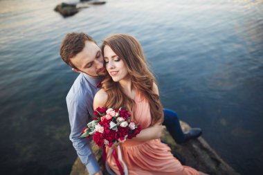 Düğün çifti mavi denizin yanındaki kayalarda öpüşüyor ve sarılıyor.