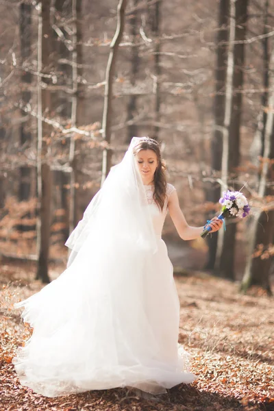 Великолепная невеста в элегантном платье держа букет и позируя рядом с лесом — стоковое фото