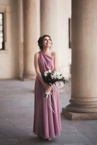 Retrato de una hermosa novia joven que se encuentra entre las columnas cerca del edificio viejo y la celebración de ramo de novia — Foto de Stock