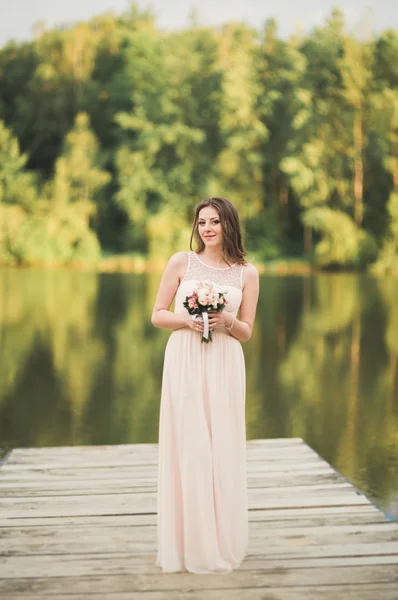 Hermosa novia en vestido elegante celebración de ramo posando cerca del bosque y el lago —  Fotos de Stock