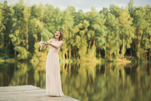 Vacker brud i elegant klänning holding bukett poserar nära skog och sjö — Stockfoto