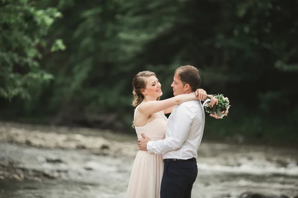 Prachtig bruidspaar zoenen en omhelzen bij de oever van een bergrivier met stenen — Stockfoto