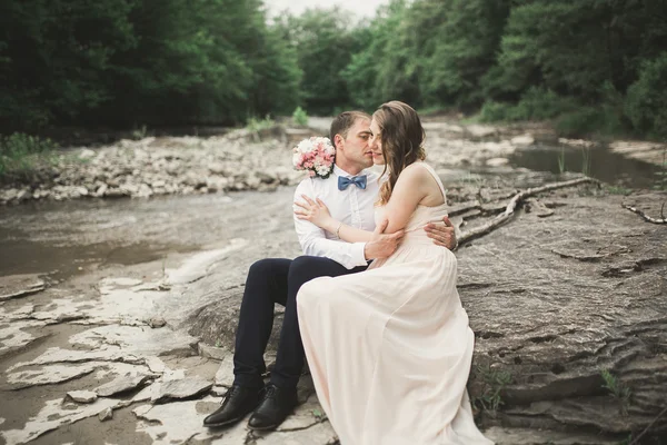 Elegante novio elegante y elegante novia cerca del río con piedras. Pareja de boda enamorada —  Fotos de Stock