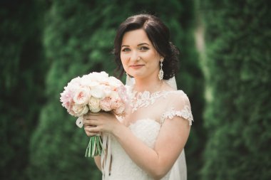 Zarif beyaz elbiseli güzel esmer gelin elinde buket ile güzel ağaçlar poz veriyor.