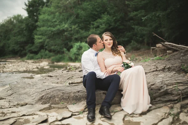 Hermosa pareja de boda besándose y abrazándose cerca de la orilla de un río de montaña con piedras —  Fotos de Stock