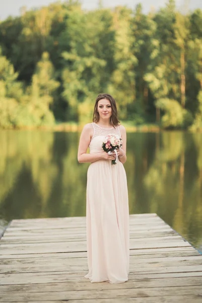 Hermosa novia en vestido elegante celebración de ramo posando cerca del bosque y el lago —  Fotos de Stock