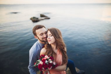 Düğün çifti mavi denizin yanındaki kayalarda öpüşüyor ve sarılıyor.