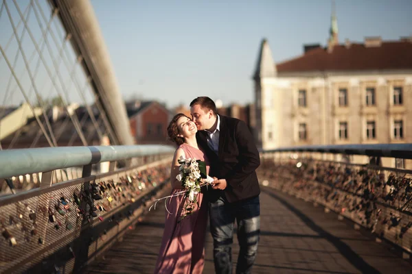 Snygg kärleksfull bröllopsparet, brudgummen, brud med rosa klänning kyssas och kramas på en bro vid solnedgången — Stockfoto