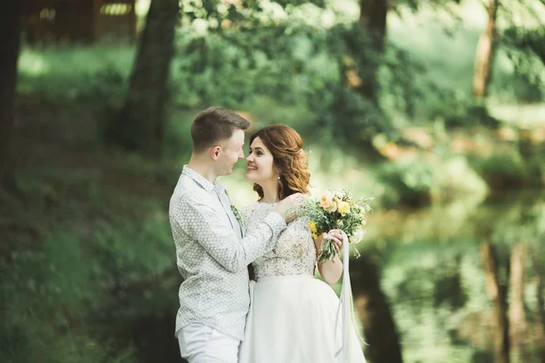 Elegante coppia di sposi felici che passeggiano nel parco il giorno del loro matrimonio con bouquet — Foto Stock