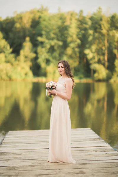 Vacker brud i elegant klänning holding bukett poserar nära skog och sjö — Stockfoto