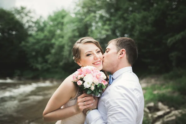 Elegante stilvolle glückliche brünette Braut und wunderschöner Bräutigam vor dem Hintergrund eines wunderschönen Wasserfalls in den Bergen — Stockfoto