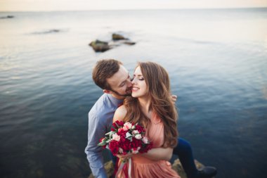 Düğün çifti mavi denizin yanındaki kayalarda öpüşüyor ve sarılıyor.