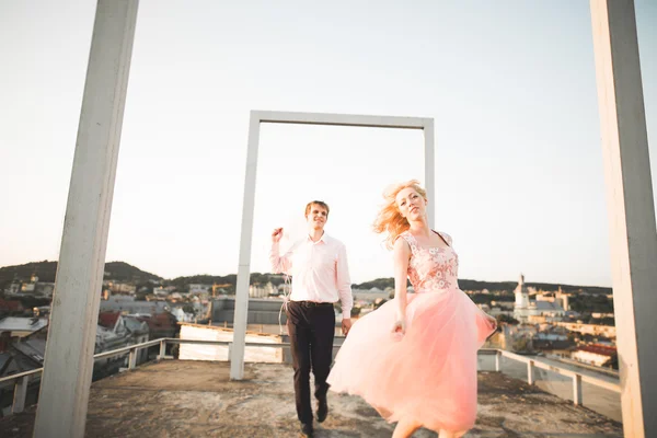 Moda hermosa pareja posando en el techo con fondo de la ciudad. Joven hombre y sensual rubia al aire libre. Estilo de vida . —  Fotos de Stock