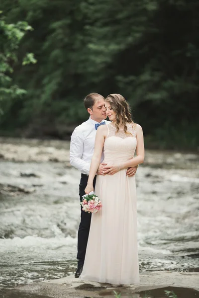 Hermosa pareja de boda besándose y abrazándose cerca de la orilla de un río de montaña con piedras —  Fotos de Stock