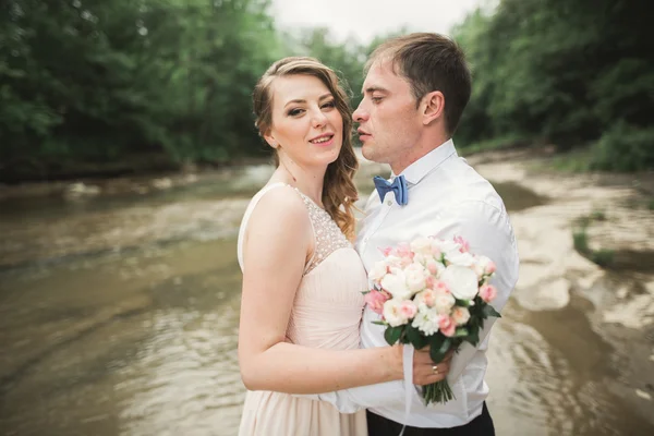 Hermosa pareja de boda besándose y abrazándose cerca de la orilla de un río de montaña con piedras — Foto de Stock