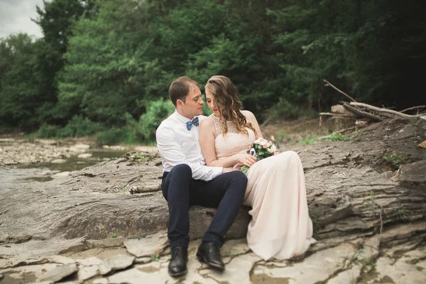 Elegante novio elegante y elegante novia cerca del río con piedras. Pareja de boda enamorada —  Fotos de Stock