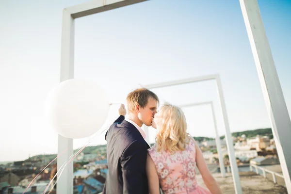 Retrato ao ar livre muito ensolarado de jovem casal elegante enquanto se beija no telhado com vista para a cidade — Fotografia de Stock