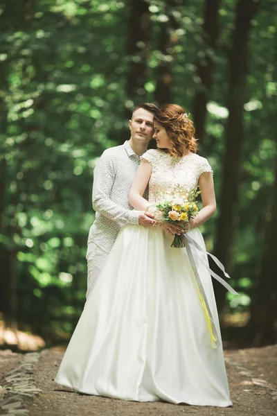 Elegante coppia di sposi felici che passeggiano nel parco il giorno del loro matrimonio con bouquet — Foto Stock