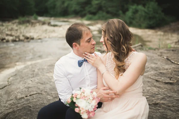 Hermosa pareja de boda besándose y abrazándose cerca de la orilla de un río de montaña con piedras —  Fotos de Stock