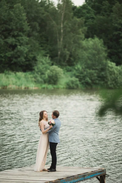 คู่แต่งงานที่สวยงามหรูหรา โพสใกล้ทะเลสาบตอนพระอาทิตย์ตก — ภาพถ่ายสต็อก