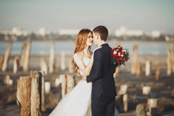 Casal de casamento jovem bonito, noiva e noivo posando perto de postes de madeira no fundo do mar — Fotografia de Stock