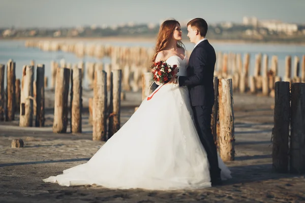 Красивая молодая супружеская пара, невеста и жених позируют возле деревянных столбов на фоне моря — стоковое фото