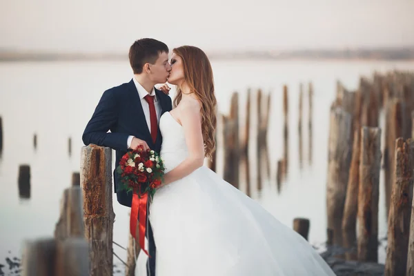 Casal de casamento jovem bonito, noiva e noivo posando perto de postes de madeira no fundo do mar — Fotografia de Stock