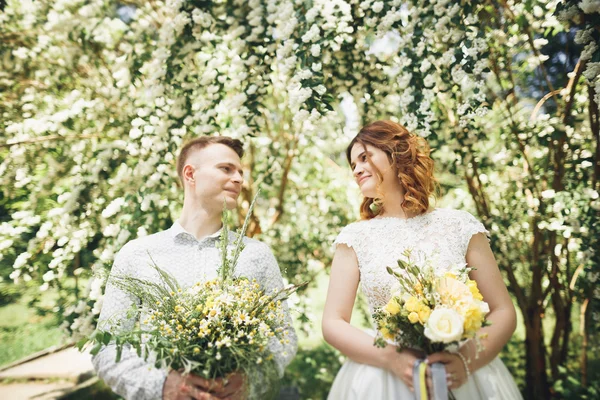 Couple élégant de jeunes mariés heureux marchant dans le parc le jour de leur mariage avec bouquet — Photo