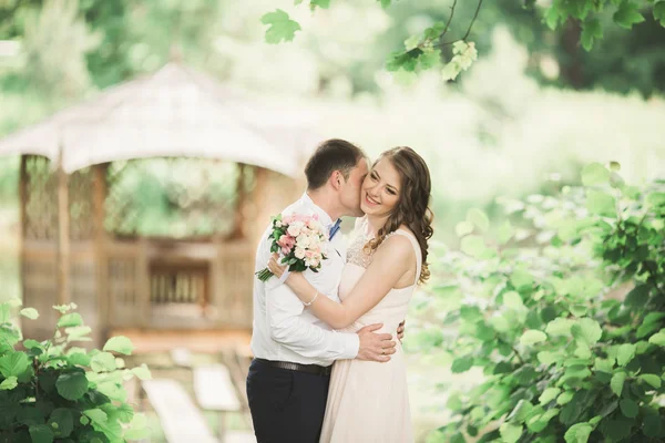 Adorável casal apaixonado beijando uns aos outros no dia do casamento, de pé no parque ao ar livre perto do lago — Fotografia de Stock