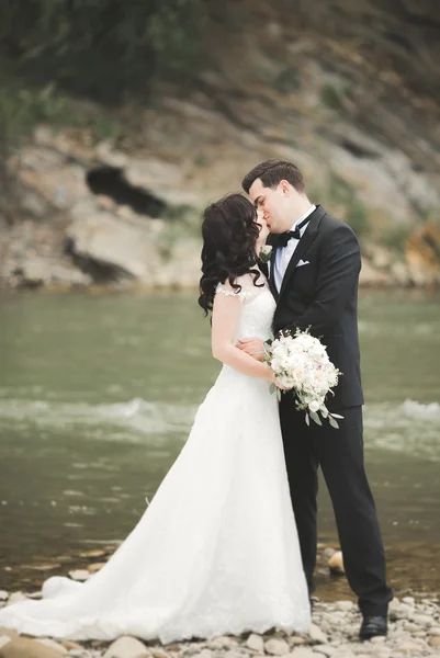 Schönes Hochzeitspaar küsst und umarmt sich am Ufer eines Gebirgsflusses mit Steinen — Stockfoto