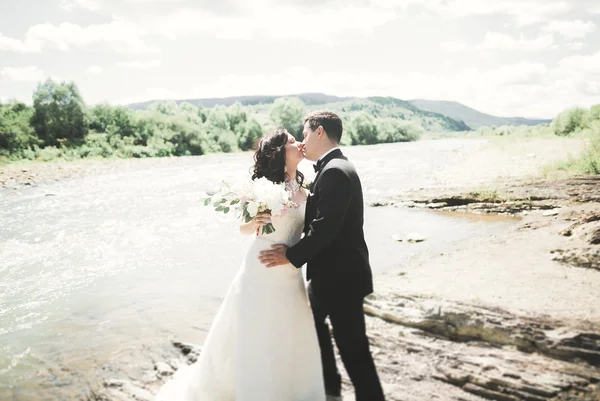 Elegante stilvolle glückliche brünette Braut und wunderschöner Bräutigam vor dem Hintergrund eines wunderschönen Wasserfalls in den Bergen — Stockfoto
