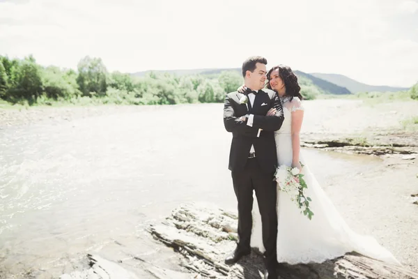 Schönes Hochzeitspaar küsst und umarmt sich am Ufer eines Gebirgsflusses mit Steinen — Stockfoto