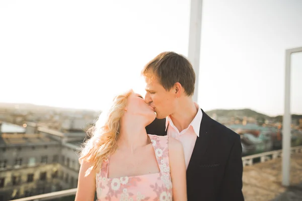 Giovane coppia innamorata posa sul tetto con perfetta vista sulla città che si tiene per mano e abbraccia. Bellissimo tramonto — Foto Stock