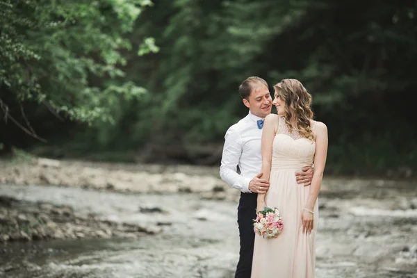 Hermosa pareja de boda besándose y abrazándose cerca de la orilla de un río de montaña con piedras —  Fotos de Stock