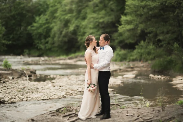 Hermosa pareja de boda besándose y abrazándose cerca de la orilla de un río de montaña con piedras —  Fotos de Stock