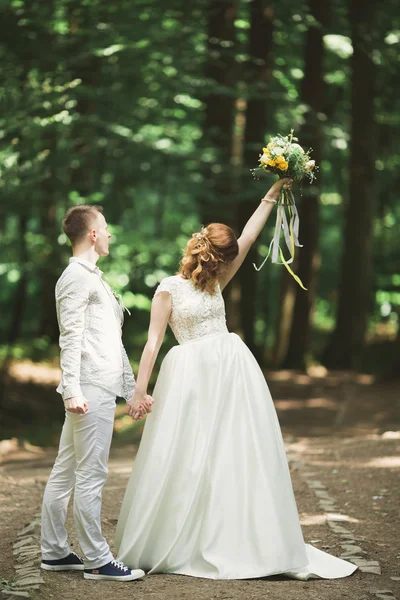 Couple élégant de jeunes mariés heureux marchant dans le parc le jour de leur mariage avec bouquet — Photo