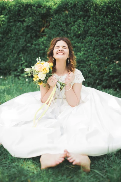 Прекрасная невеста сидит на земле, держа букет, улыбаясь в камеру — стоковое фото