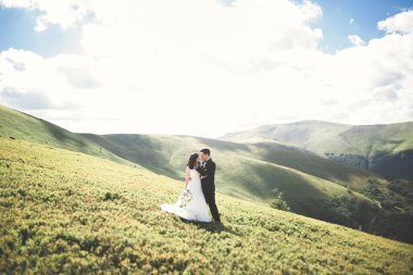 Güzel, mutlu gelin ve şık damat güneşli, çarpıcı dağların arka planında poz veriyor.