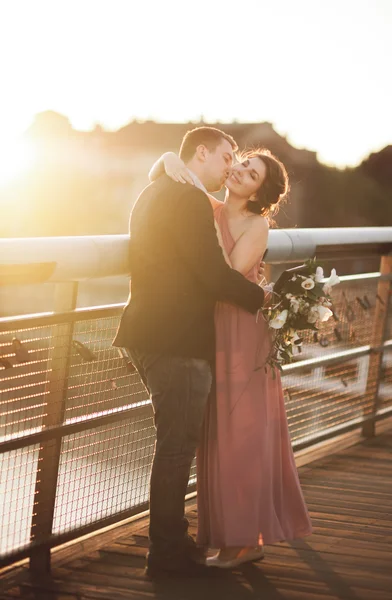 Elegáns szerelmes esküvő házaspár, vőlegény, menyasszony, rózsaszín ruhát csókolózás, és átölelte a hídon naplementekor — Stock Fotó