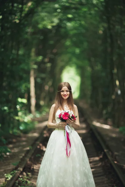 Hermosa novia morena en elegante vestido blanco sosteniendo ramo posando árboles limpios — Foto de Stock