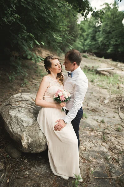 Hermosa pareja de boda besándose y abrazándose cerca de la orilla de un río de montaña con piedras —  Fotos de Stock